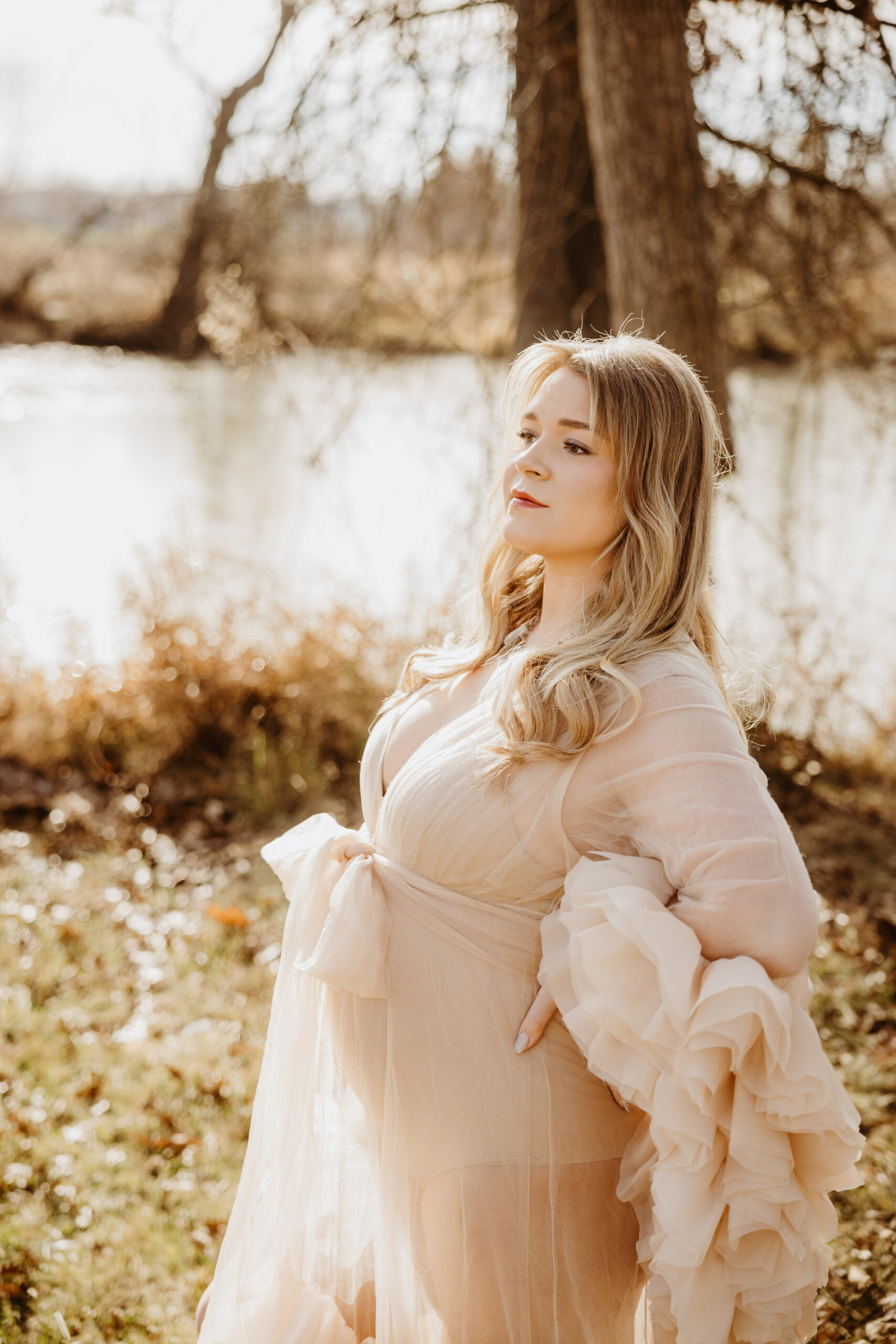 Pregnant woman holds belly in flowing dress in front of lake